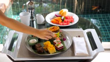 Indulgent breakfast on luxury villa terrace. Woman chief decorate a cake with slices of fresh strawberry. Table filled with tropical fruits, desserts, and cakes. Served breakfast on tropical resort.