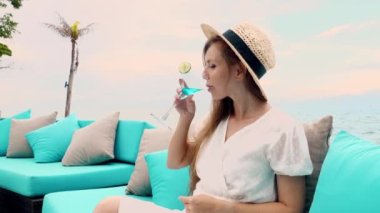 Young woman in a straw hat lounges on a beach couch in island, while enjoying a refreshing cocktail. Happy travel girl on tropical summer vacation.