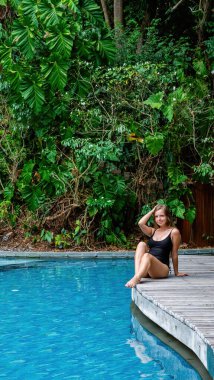 Tan young model in black bikini, posing and relaxing by swimming pool. Tropical island vacation.