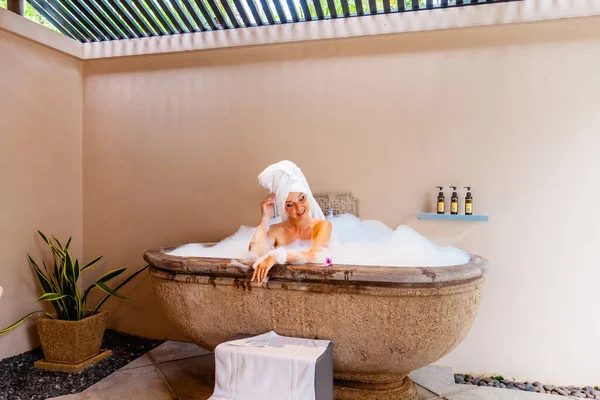 Outdoor bath at luxurious spa hotel. A beautiful young woman relaxes in a foam filled tub, with exotic flowers and organic skin care products. Self-care and spa concepts, inspire relaxation.