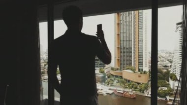 Blogger in hotel room with city view, walking to window while talking on phone, showing his view on social media. Influencer concept. Slow motion. Megacity river view.