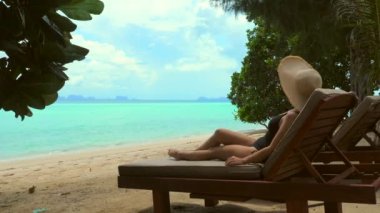 Young woman on vacation enjoying sea view in luxury hotel by the beach. Travel girl in bikini and hat, relaxing on a sunbed. The perfect tropical escape for a summer holiday.