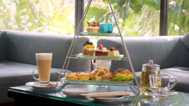 Afternoon tea stand with colorful macarons, petit cakes, and tiny sandwiches, paired with a glass teapot and cup of coffee with tropical palm trees on background. Delicious sweet food.
