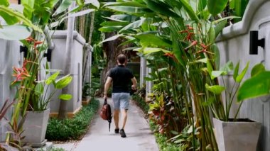 Male traveler with a backpack in his hands walking on villa in modern luxury tropical resort. Hotel in jungle. A man walks through the hotel among the greenery and trees. Back view