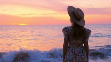 Cennet sahilindeki altın pembe gün batımındaki kadının arka görüntüsü. Tayland 'da yaz tatilindeki kadın turistin yavaş çekimde uzaklaşması. Okyanus Akdeniz seyahatinde el sallayan kadın elbisesi.