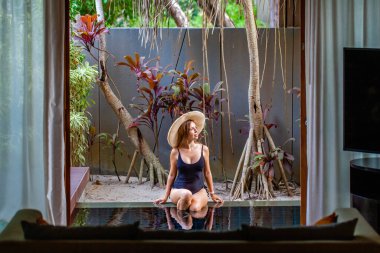 Travel woman in straw hat and black swimsuit sitting on pool edge on tropical luxury villa in modern resort. Summer tropical vacation in Thailand. 