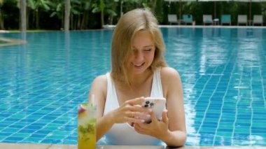 Happy smiling woman with mobile phone drinking tropical cocktail in swimming pool at luxury resort. Girl relaxing sunbathing enjoy travel holidays using smart phone surfing social media, booking hotel