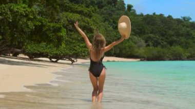 Tropikal plajda zıplayan ve dans eden neşeli kadın, tropikal tatilin tadını çıkar. Kadın turist güneşli bir günde arka planda turkuaz deniz manzarasıyla eğleniyor. Arkadan bak. Yavaş çekim