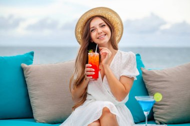 Hasır şapkalı genç romantik bir kadın açık hava barında alkolik mavi lagün ve mai tai kokteylleriyle masaya oturur. Deniz altyapısı, lüks otellerde restoran, otel. Rahatla, dinlen, tatil, tatil