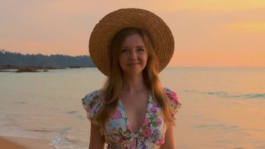Kameraya bakıp gülümseyen çekici genç bir kadının portresi tropikal gün batımı deniz sahilinde duruyor, yavaş çekim yakın görüntü. Yaz tatili. Büyüt.