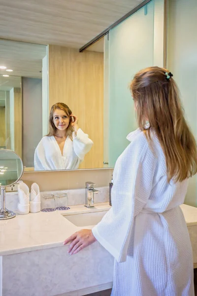 迷人的年轻女人穿着浴衣 在浴室的镜子里涂上面霜 欣赏自己健康的皮肤 护肤和化妆概念 用于晨间常规治疗或温泉治疗 — 图库照片