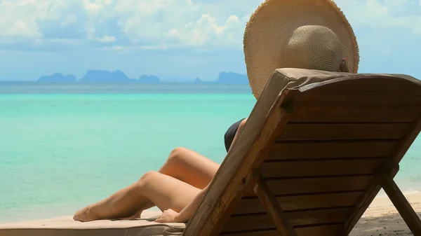 Unrecognizable young woman in a bikini and hat relaxing on a comfortable lounger beach chair, overlooking the turquoise waters of a tropical paradise. Summer tropical getaway.