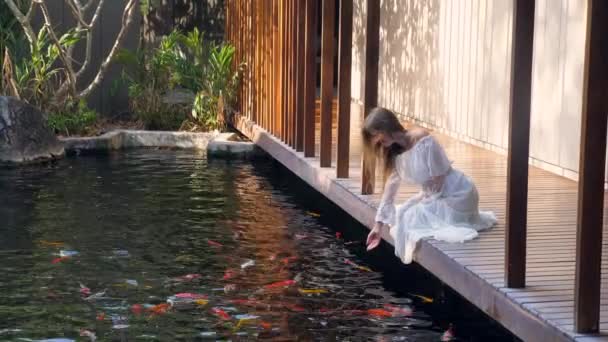 身着白衣的女人在有鱼塘的豪华温泉放松的慢镜头 坐在木桥上欣赏大自然 — 图库视频影像