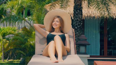 Yaz tatilinde açık havanın tadını çıkaran çekici bir kadın, plaj sandalyesinde okyanus kenarına uzanmış, palmiye ağaçlarıyla çevrili. Gülümseyen kadın gezgin, deniz kenarında güneşlenen..
