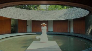 Beyaz elbiseli bir kadın lüks bir otel ya da tatil köyünde güzel yeşil bir göletle çevrili bir spa kompleksinde dinleniyor. Seyahat, Asya tatili. Yavaş çekim.