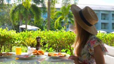 Tayland 'da güneşli bir yaz gününde, elbiseli çarpıcı bir kadın açık hava restoranında rahatlatıcı bir yemek yiyor. Yaz havası, tropikal tatil kahvaltısı. Yan görünüm.