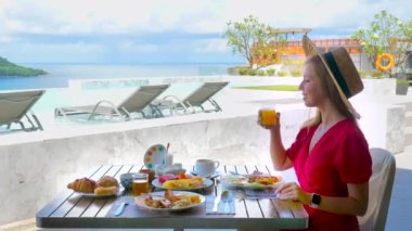 Hasır şapkalı gezgin kız, deniz manzaralı lüks otel otelinde kahvaltıda portakal suyu içiyor. Yaz tatilindeki genç bir kadın cennete gider. Tropik tatil. Yan görünüm.