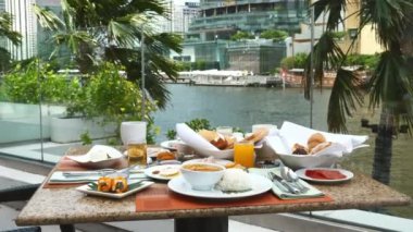 Açık hava lüks otel restoranında taze hamur işi, jambon, sosis, peynir ve meyve suyu. Arka planda tekneler var. Nehir manzaralı büfe otel restoranı. Tropik şehir tatili, tatil.