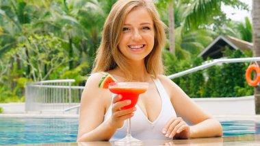 Beautiful woman in white swimsuit relaxing, enjoying cocktail by outdoor pool. Happy, smiling and having fun. Travel, vacation, tropical holidays.