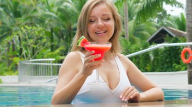 Beautiful woman in swimsuit with cocktail at outdoor swimming pool. Female traveler with smile on her face, drink summer tropical cocktail, looking at camera, creating happy and carefree atmosphere