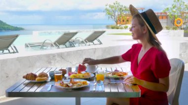 Yaz tatilindeki genç kadın, deniz manzaralı lüks otel otelinde tropikal meyvelerin tadını çıkarıyor. Hasır şapkalı mutlu turist kahvaltıda masada oturuyor..