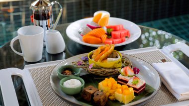 Tropical desserts, cakes, and fruits on a table set with plates. Slices of cakes on a white tray. Mango, chocolate, red velvet, sandwiches.
