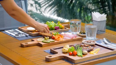Restoran garsonu açık hava tropikal restoranında yemek servisi yapıyor. Garson masaya ahşap bir yemek tahtası koyuyor. Restoran personeli güzel bir yemek hazırlar..