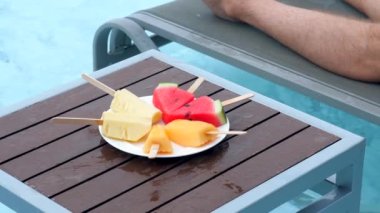 Genç adam havuzda karpuz, ananas ve karpuz ile eğleniyor. Lüks havuz başı partisinde yaz tatilinin tadını çıkaran bir erkeğin yavaş çekim görüntüleri..