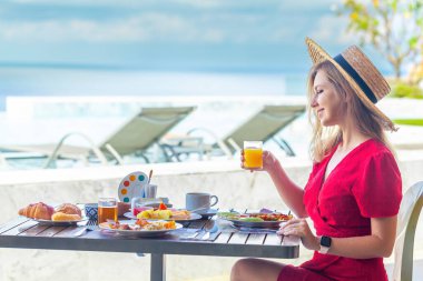 Yaz tatilinde olan genç bir kadın tropikal meyvelerle kahvaltı yapıyor, deniz manzaralı lüks bir otel terasında portakal suyu içiyor..