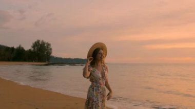 Gün batımında tropik deniz sahillerinde yürüyen ve eğlenen mutlu ve çekici genç bir kadının yavaş çekimi. Dişi gezgin gün batımında hayatın tadını çıkarır. Seyahat, yaz tatili.