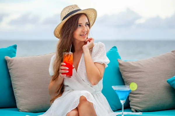 stock image Young romantic woman in straw hat sits by table with alcoholic blue lagoon and mai tai cocktails at outdoors bar with sea background, restaurant at luxury resort, hotel. Relax, rest, holiday, vacation