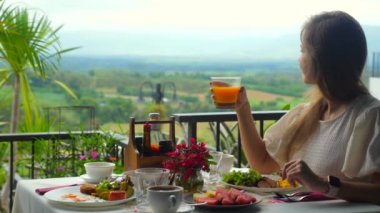 Gezgin lezzetli kahvaltı yemekleriyle dolu masada oturarak ferahlatıcı bir bardak portakal suyu içmeyi sever. Arka planda yeşil vadi manzarası, tatil beldesinde yaz tatili. Yaz gezisi kavramı, tatil