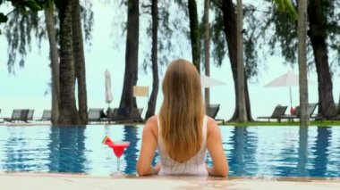 Back view of woman model, lounging in blue swimming pool in white bikini, tan during summer vacation. Relaxation at spa resort. Woman enjoying perfect beach holiday vacation at luxury beachfront hotel