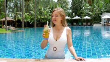 Genç bir kadın tropik cennette güneşlenirken havuzun kenarında kokteyl yudumluyor. Palmiye ağaçları bu rahatlatıcı tatil sahnesi için pitoresk bir arkaplan sağlar.