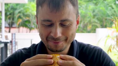 Adam açık hava restoranında sulu hamburger yiyor. İnsan lezzetli ve sağlıksız yemekten hoşlanır. Komik ve duygusal ifadeler kullanır. Büyük burgeri iki eliyle tutar ve büyük lokma yer.