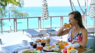 Kahvaltıda ya da kahvaltıda portakal suyu içen mutlu genç bir kadın. Güneşli yaz gününde yemek, meyve, pasta ve kahve dolu bir masada. Seyahat, tatil, tatil, hayatın tadını çıkar. Mavi deniz arka planı.