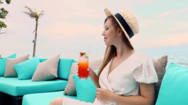 Young tourist girl in a straw hat relaxes on a beach couch, enjoying a alcoholic cocktail and taking in the beautiful sea view. Holidays and summer vacations.