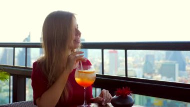 Modern Bangkok 'ta yüksek çatı barının ya da restoran terasının kenarında oturan kokteyl içen genç ve mutlu bir kadın. Akşam istirahatı, işten sonra dinlenme. Arkaplanda gökdelenler.
