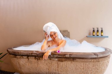 Outdoor bath at luxurious spa hotel. A beautiful young woman relaxes in a foam filled tub, with exotic flowers and organic skin care products. Self-care and spa concepts, inspire relaxation.