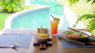 Açık havuz kenarındaki bir kafede bir garson masaya yeni hazırlanmış bir tabak ve içecekler koyar. Tropik bir kafede havuz manzaralı güzel bir hamburger servisi. Tatil yemeği.