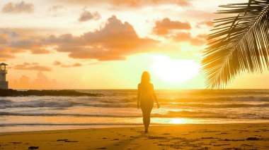 Young woman enjoy summer vacation holidays at ocean. Happy female tourist walk in romantic golden sunset lights with sea waves background. Girl walking barefoot at seaside on sandy beach. Slow motion.