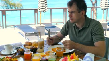Morali bozuk erkek turist sabahları tatil köyündeki kafede yemek yiyor. Üzgün düşünceli bir adam oteldeki restoranda kahvaltı yapıyor, turkuaz deniz altyapısı var. Seyahat, seyahat, tatil, tatil konsepti.
