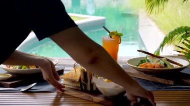Tanınmayan bir garson bir açık hava restoranında zarif bir masa hazırlıyor. Havuzu ve palmiye ağaçları olan bir öğle yemeği. Lüks ve tropikal bir ortam..
