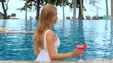 Woman luxuriating in infinity pool located at high-end hotel drinking non-alcoholic cocktail. Young woman taking pleasure in holiday in warm climate, summer vacation. Healthy lifestyle. Slow motion
