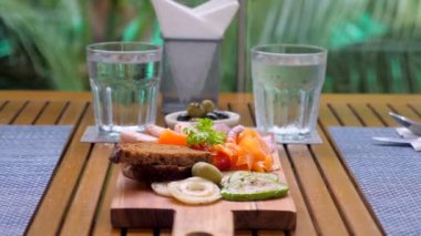 Rustik ahşap kesim tahtası füme somon, salam ve ızgara sebzeler de dahil olmak üzere çeşitli sebzeler sergiliyor. Tropik bir geçmişi olan açık bir kafede nefis bir öğle yemeği..