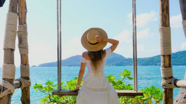 Hasır şapkalı genç bir kadın salıncakta oturup okyanus manzarasının tadını çıkarıyor. Deniz kıyısında mükemmel bir yaz tatili, dinlenme ve özgürlük anı..