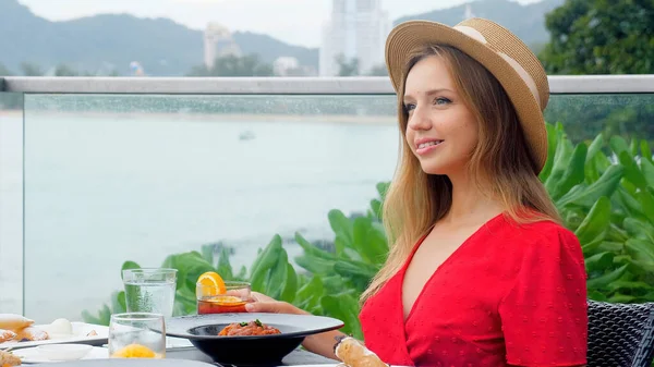 Akşam yemeğinde şapkalı ve elbiseli genç bir kadın tropik bir plajda lüks bir tatil köyünde bir bardak viskiyle. Deniz kenarındaki kafede akşam, okyanus manzaralı..