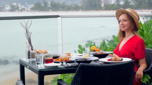 一个微笑的女人坐在热带海滩上的一家度假胜地餐厅的桌旁 年轻的女旅行家一边欣赏大海的壮丽景色 一边享受着晚餐 — 图库视频影像