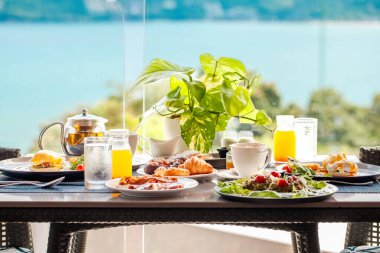 Deniz manzaralı iki kişilik açık hava otel kahvaltısı. Avokado tostu, portakal suyu, kahve, yumurta, masada servis edilen granola. Sağlıklı ve lezzetli bir yemek. Seyahat, tatil, yaz tatili.