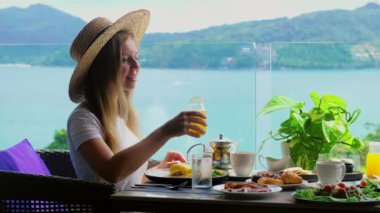Kahve masasında oturmuş, deniz manzaralı gülümseyen kadın bir bardak portakal suyu içiyor. Otelde kahvaltı. Tropik tatil beldesinde yaz tatili. Phuket Adası, Tayland, Asya. Seyahat reklamı, kahvaltı büfesi..